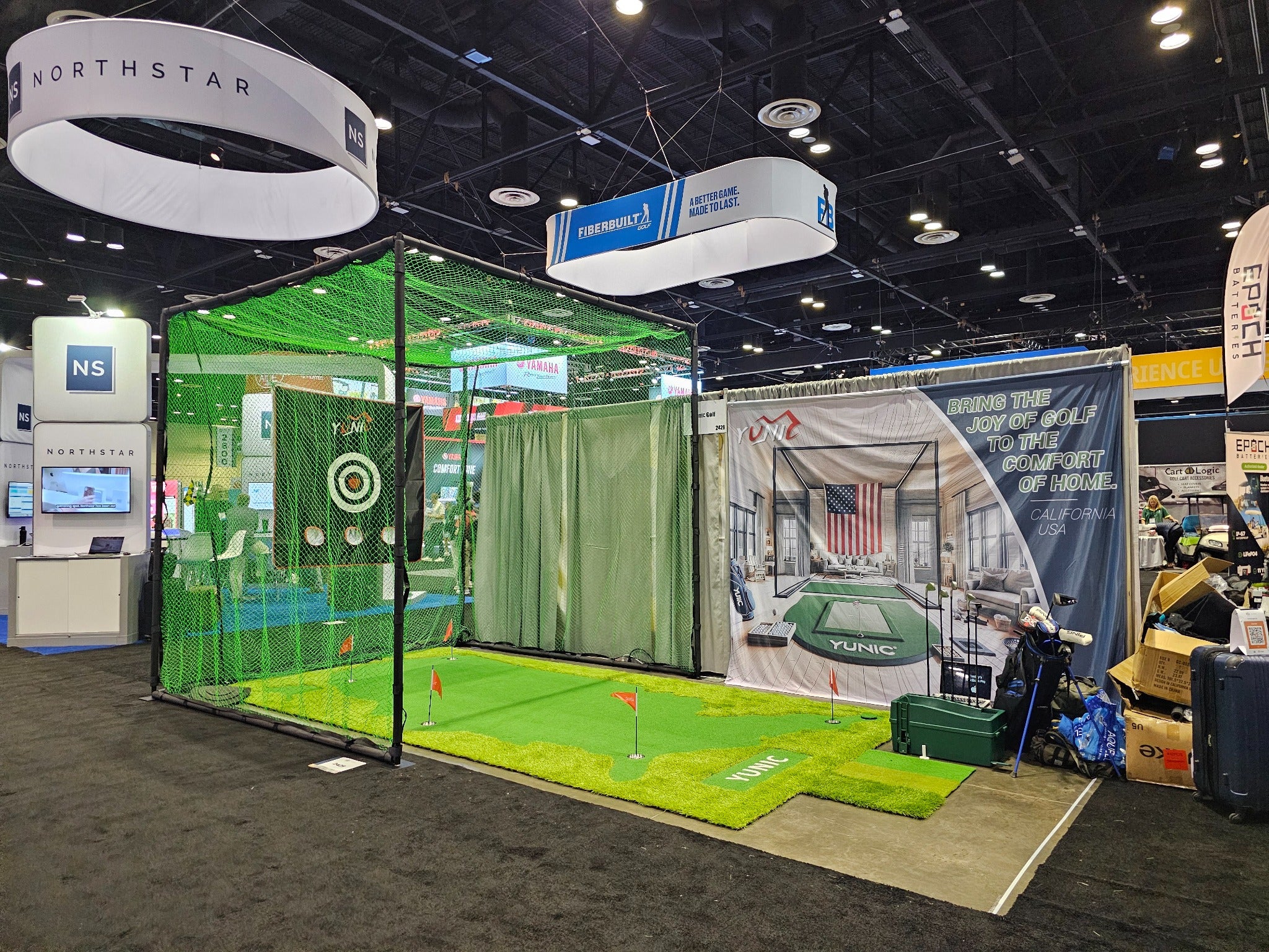YUNIC exhibit at the PGA Show, with a green-net driving cage, putting greens, and a branded booth backdrop.